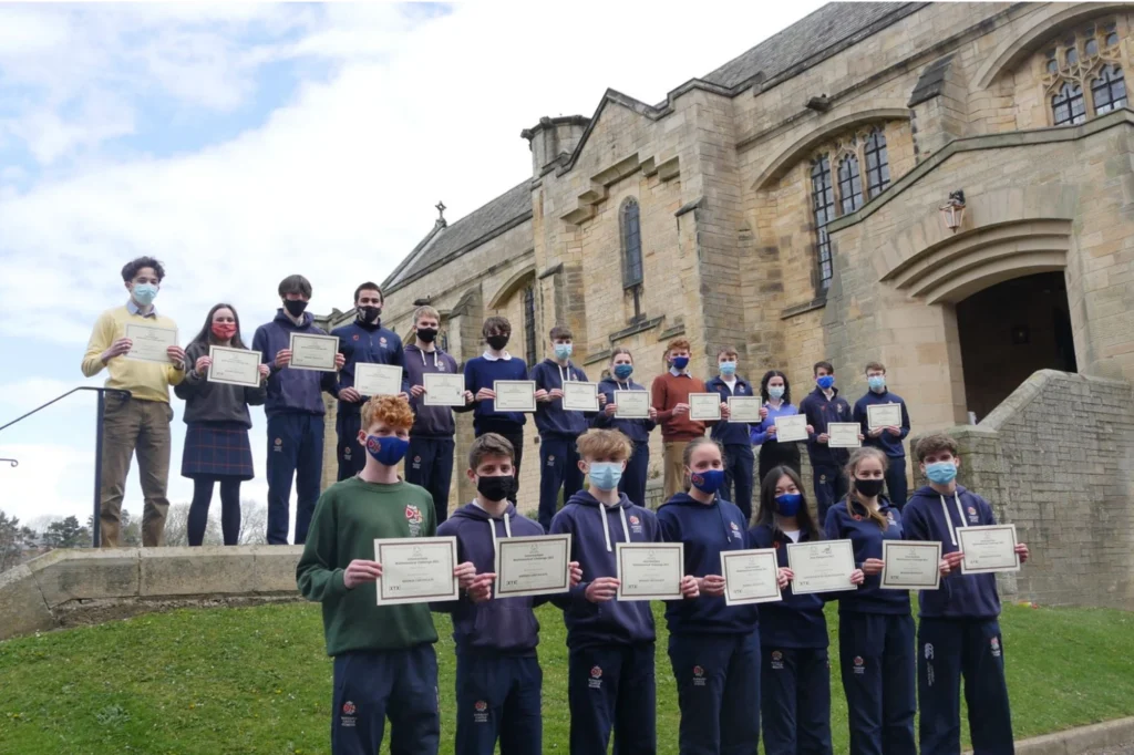 A large group of students, some wearing face masks, stand in rows proudly holding up their mathematical achievement certificates.
