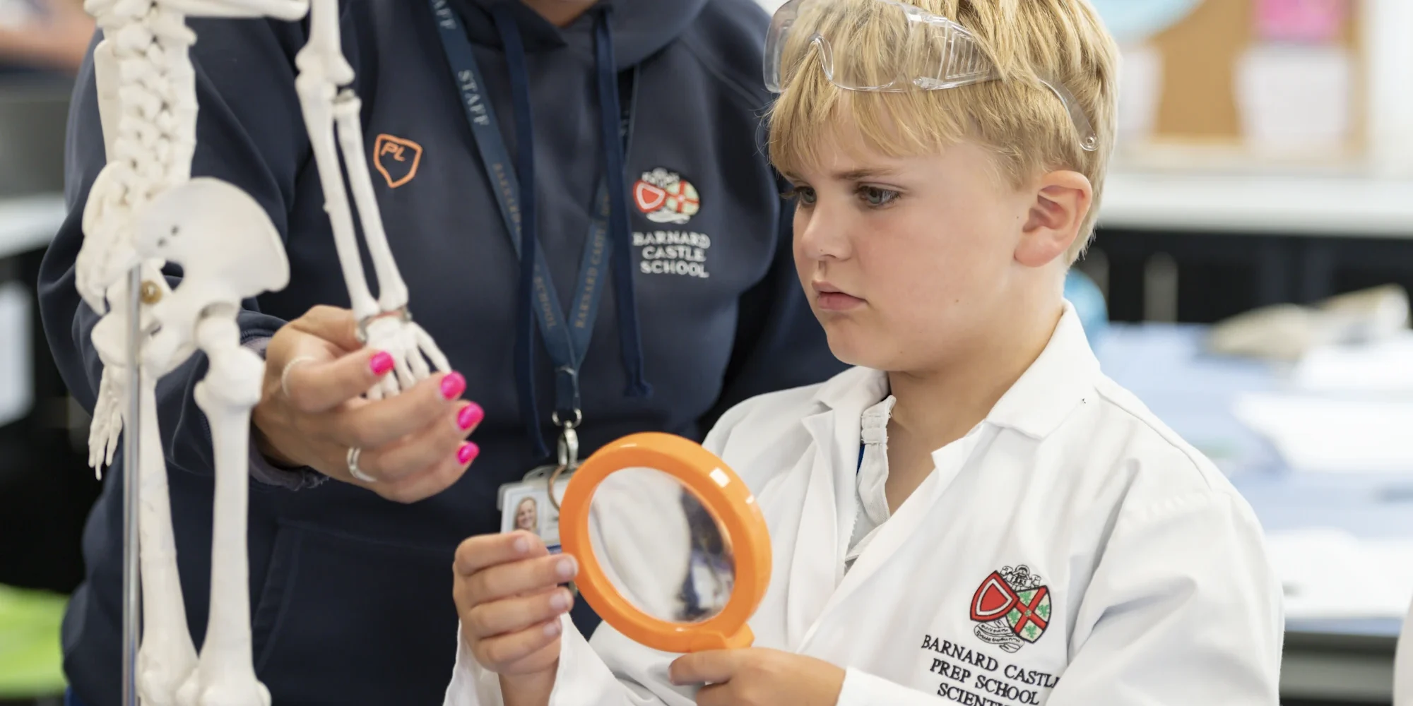 A young Barnard Castle School pupil in a lab coat, holding a magnifying glass with a teacher in the background.