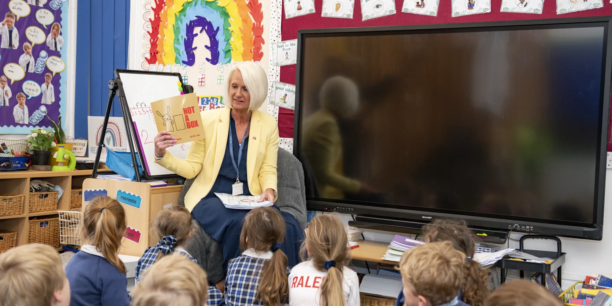A Barnard Castle School teacher holding a book, in front of a class of Preppies who are sat on the mat.
