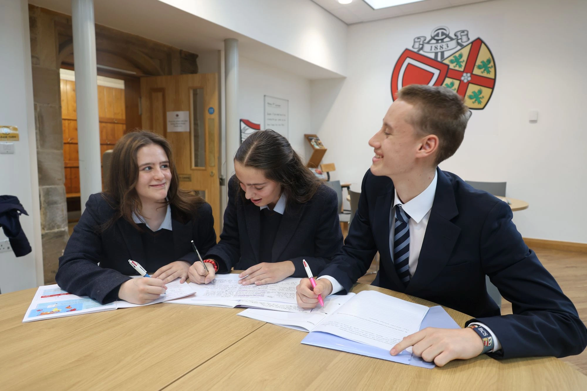 Three students in school uniforms sit at a round wooden table, smiling and writing in their notebooks.