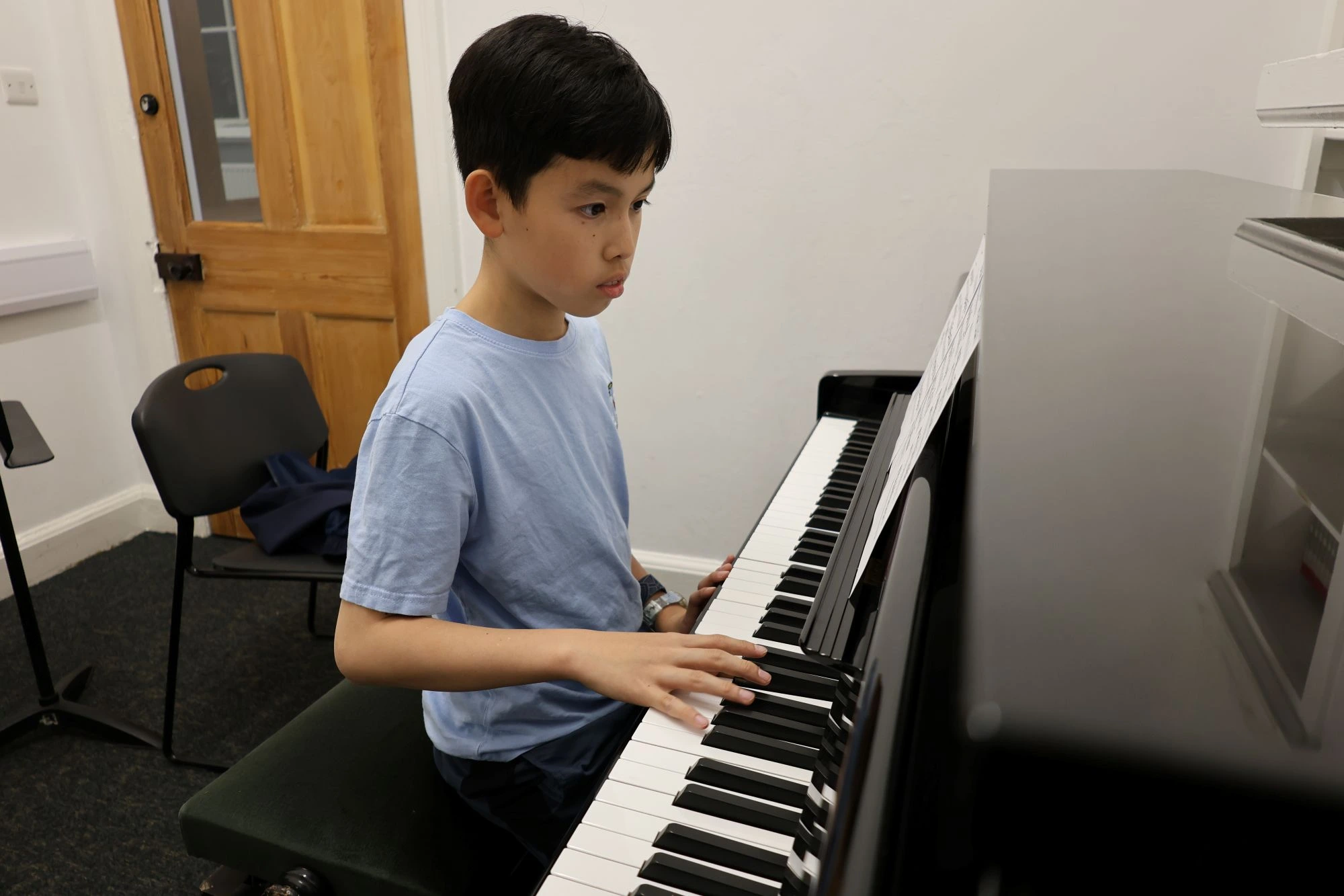 A student in a light blue t-shirt sits at a piano, focused on playing the keys in a music room.